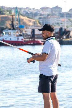 A man fishing in a harbor with industrial background on a clear day.