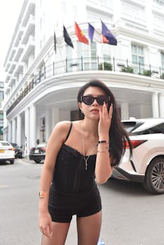 Confident woman in sunglasses posing near luxury hotel in city backdrop, day time.