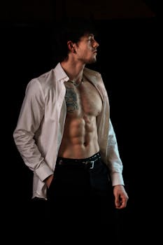 Fit man with tattoo and open shirt posing dramatically in a dark studio setting.
