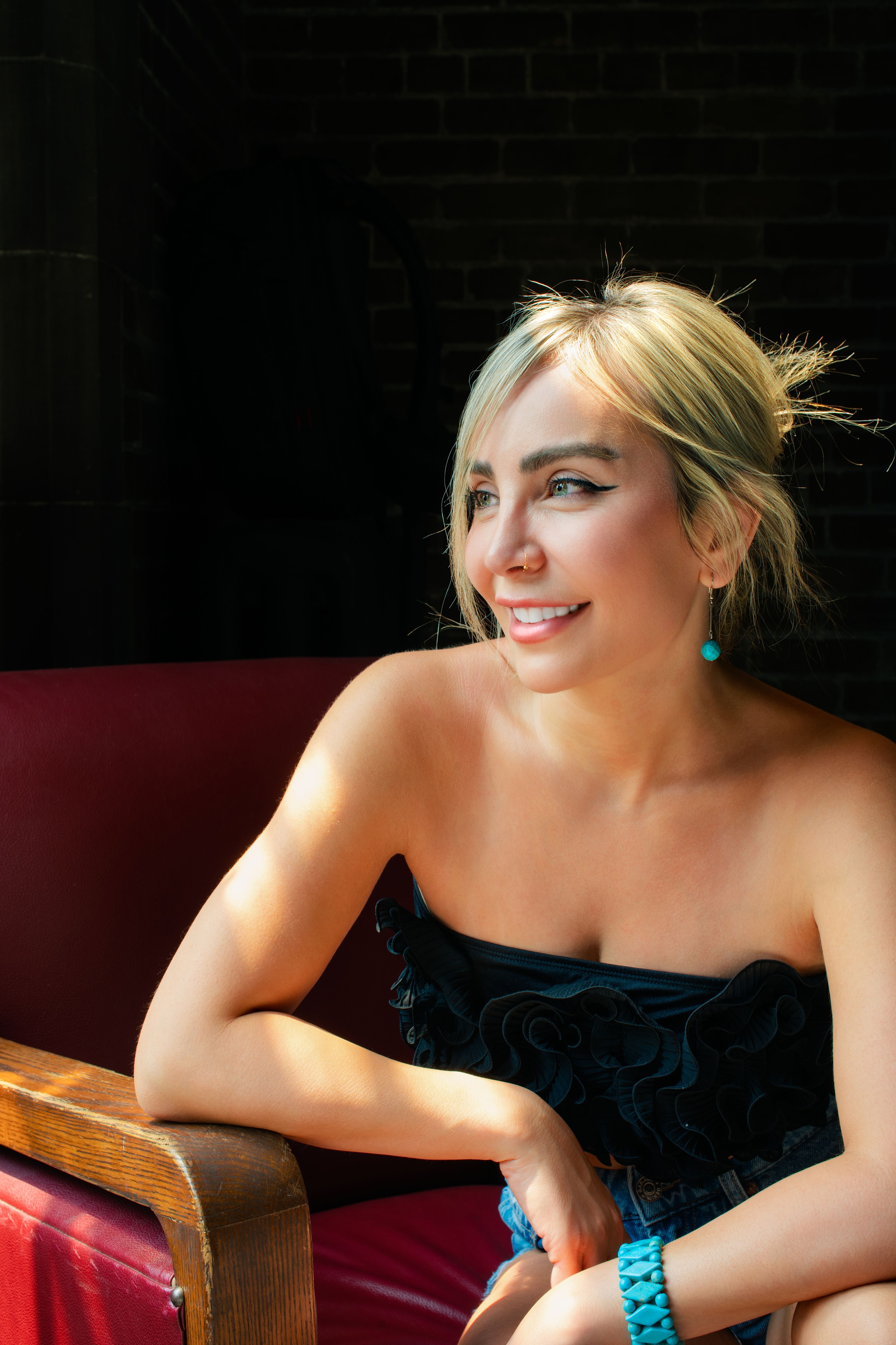 Portrait of a smiling woman sitting in a sunlit room, wearing a strapless top and turquoise accents.
