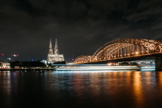 Beautiful nighttime long-exposure of Cologne Cathedral and Hohenzollern Bridge reflected over the Rhine River.