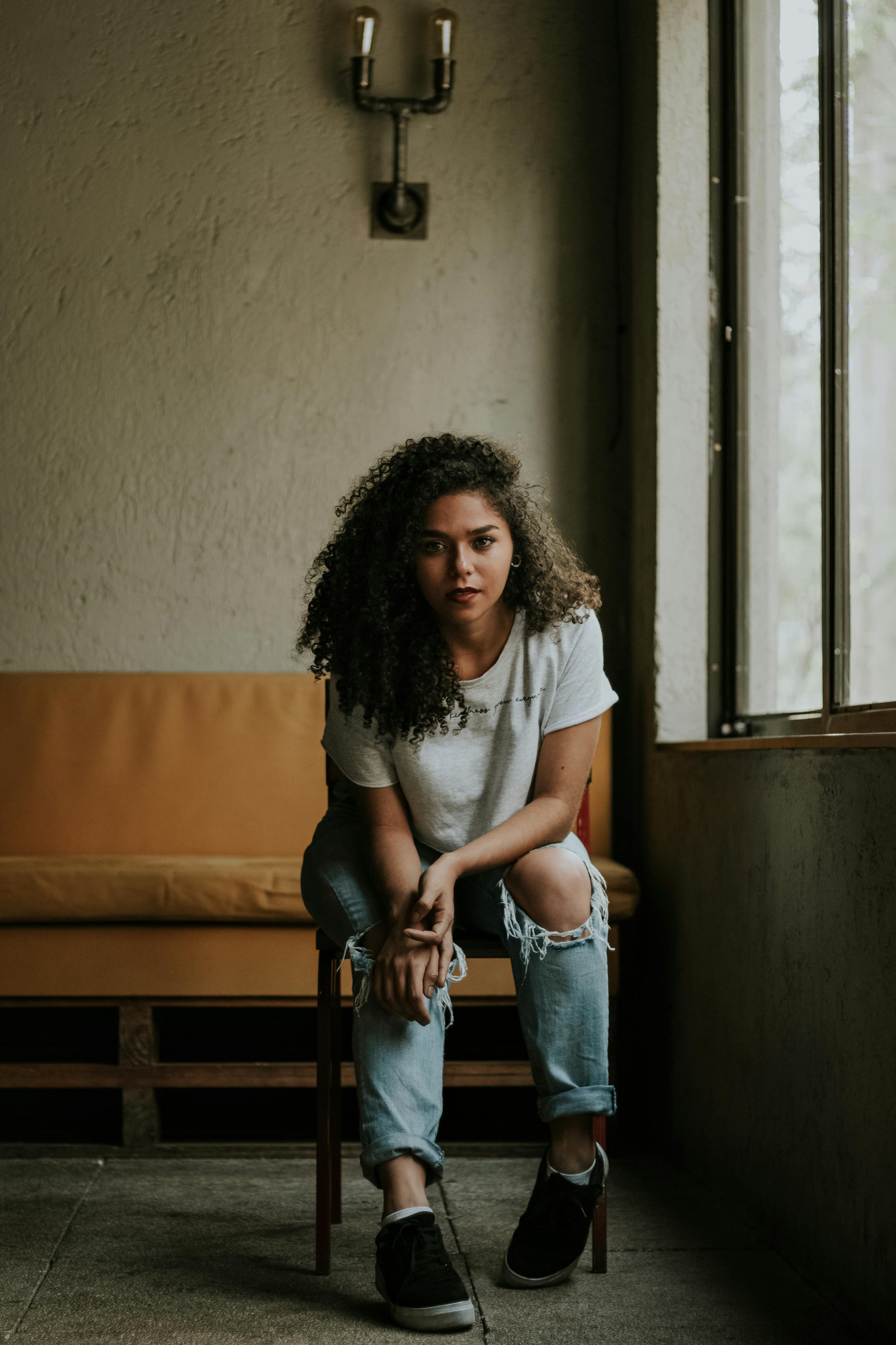 一名卷发的年轻女子穿着休闲牛仔裤和白衬衫，坐在室内的椅子上