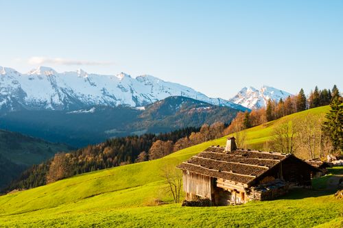Free Rustic wooden cabin nestled in lush green hills with majestic snowy mountains in the background. Perfect for nature lovers. Stock Photo