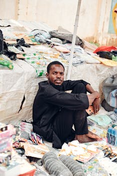 Street vendor sitting amidst diverse colorful products at outdoor market.