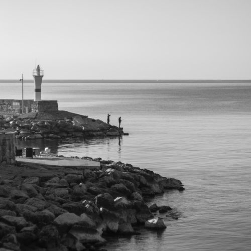 Free Serene black and white scene of a lighthouse by the coast with two people fishing. Stock Photo