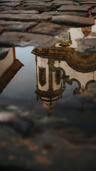 Reflection of a historic church clock tower in a rain puddle on cobblestone pavement.