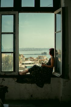 A woman sitting in a window, gazing at a scenic city view in Baku, Azerbaijan, with a serene mood.