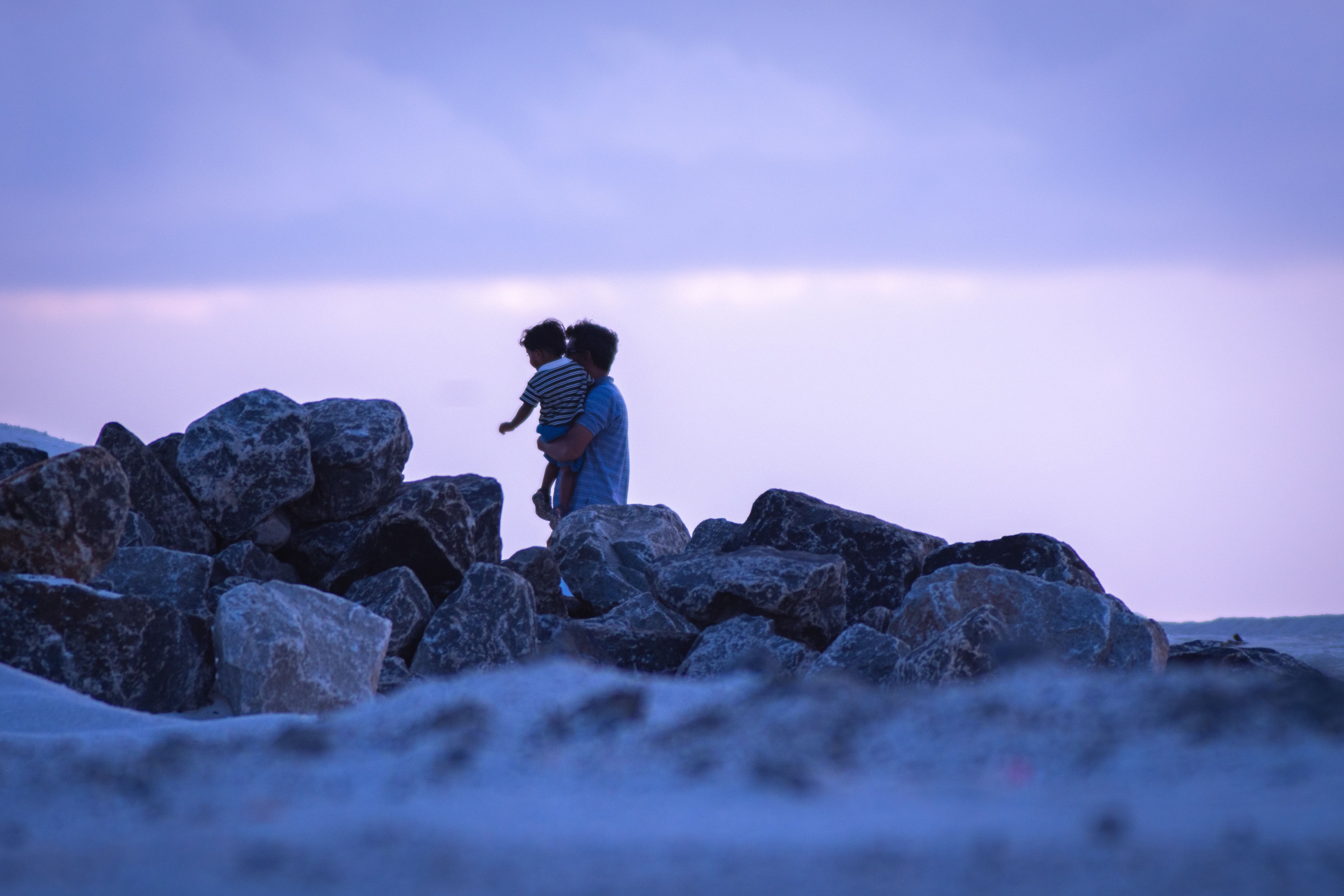 A serene scene of a father holding his child on rocky shore during twilight, capturing familial love and nature's calmness.