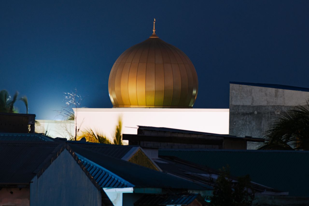 A golden mosque dome illuminated at night amidst urban rooftops, creating a serene atmosphere.