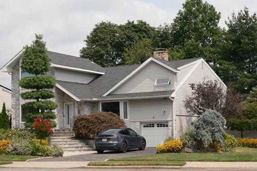 A gray two-story suburban house surrounded by lush greenery with a car parked in the driveway.