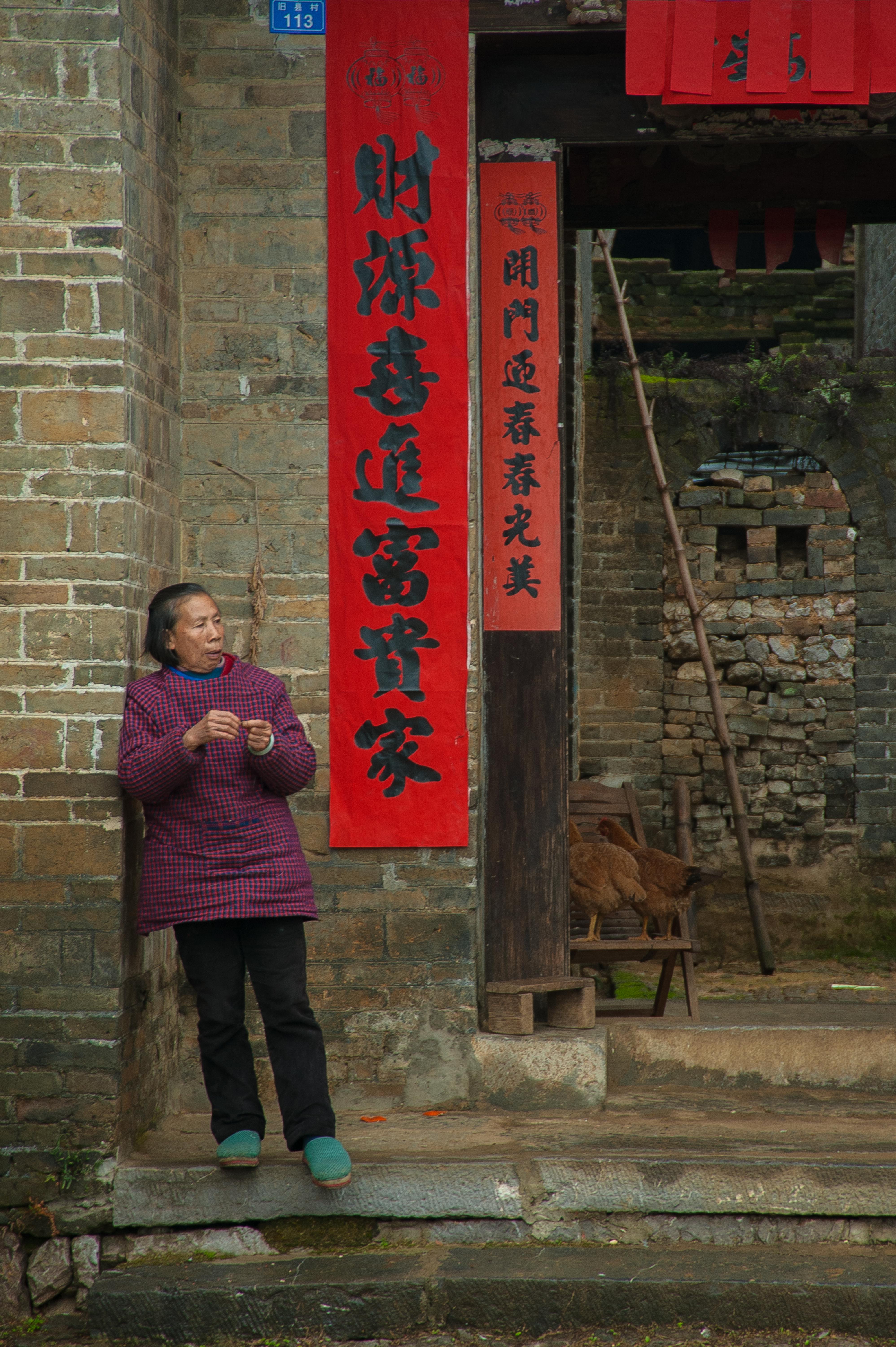An elderly woman stands by a traditional Chinese building with red banners in Guilin, China.