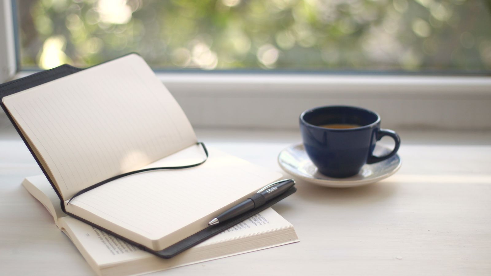 An open notebook, pen and cup beside a window