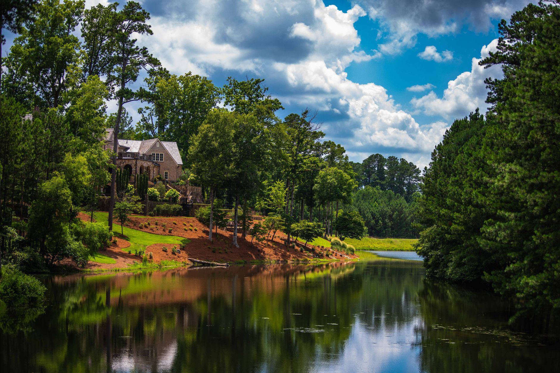 Tranquil view of a house by the forest creek in summer, reflecting lush greenery and cloudy skies.