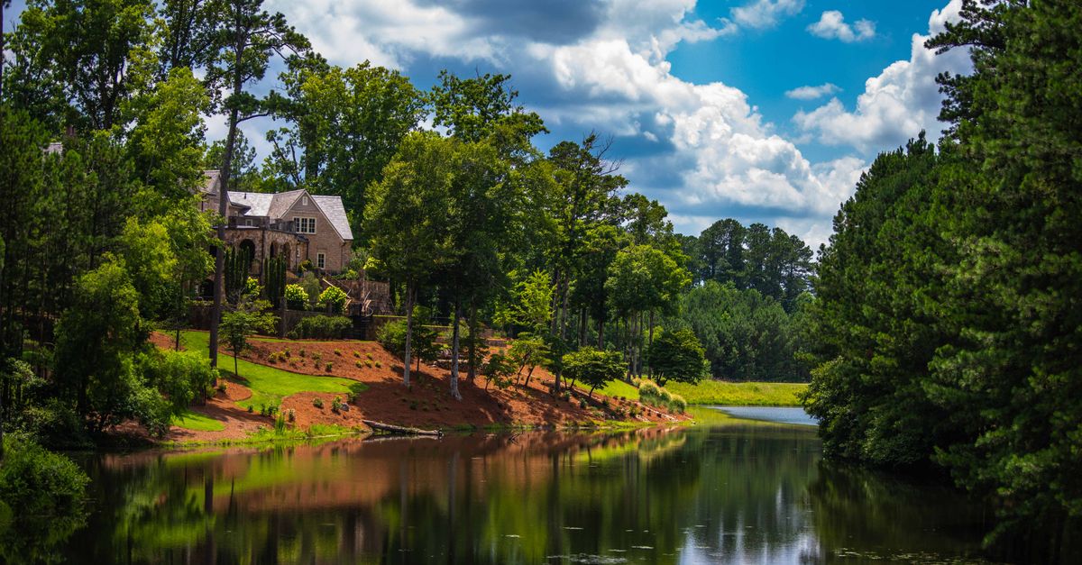 Tranquil view of a house by the forest creek in summer, reflecting lush greenery and cloudy skies.