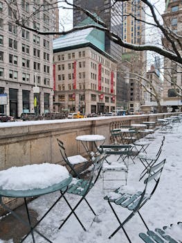 Snow-covered chairs and tables in an urban area surrounded by tall buildings during winter.