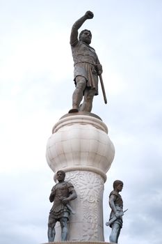 A towering warrior statue in heroic pose atop an ornate pedestal, set against a cloudy sky.