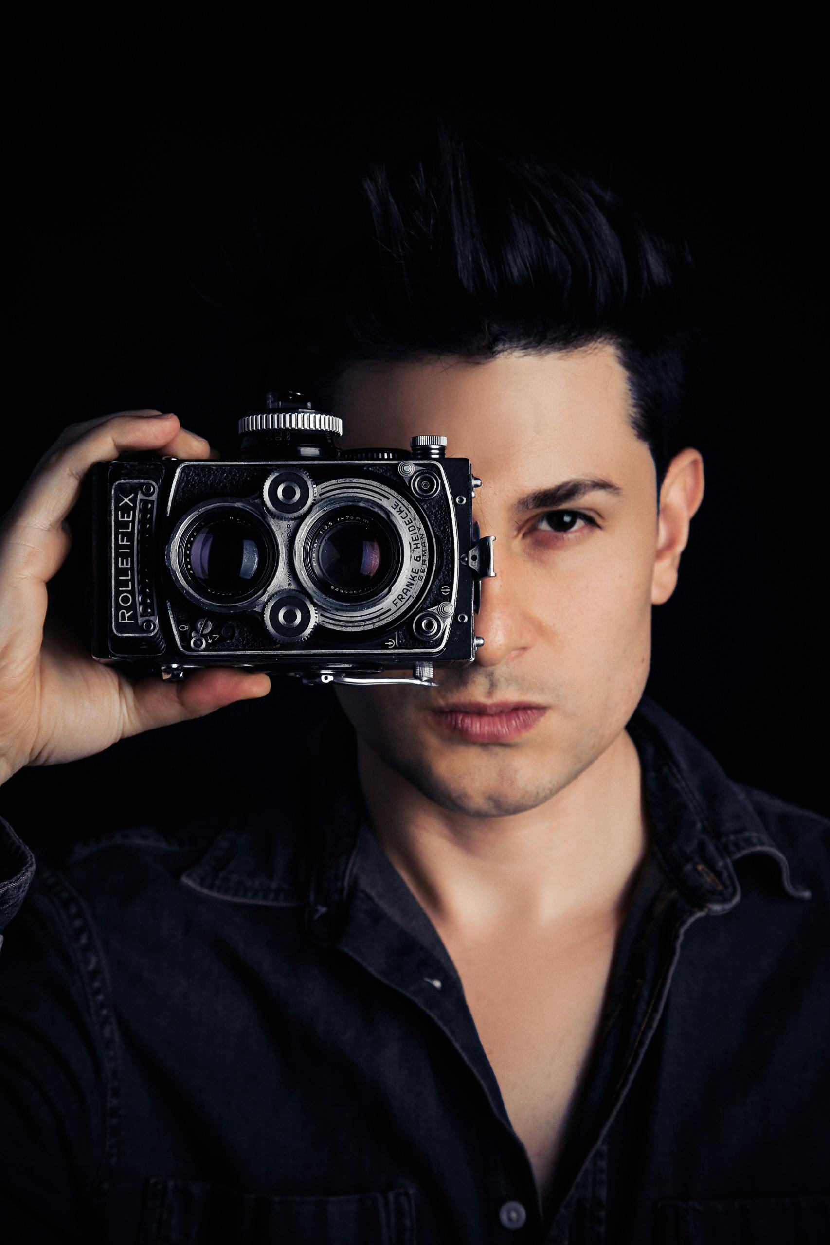 A man holding a vintage Rolleiflex camera in front of his face in a dimly lit setting.