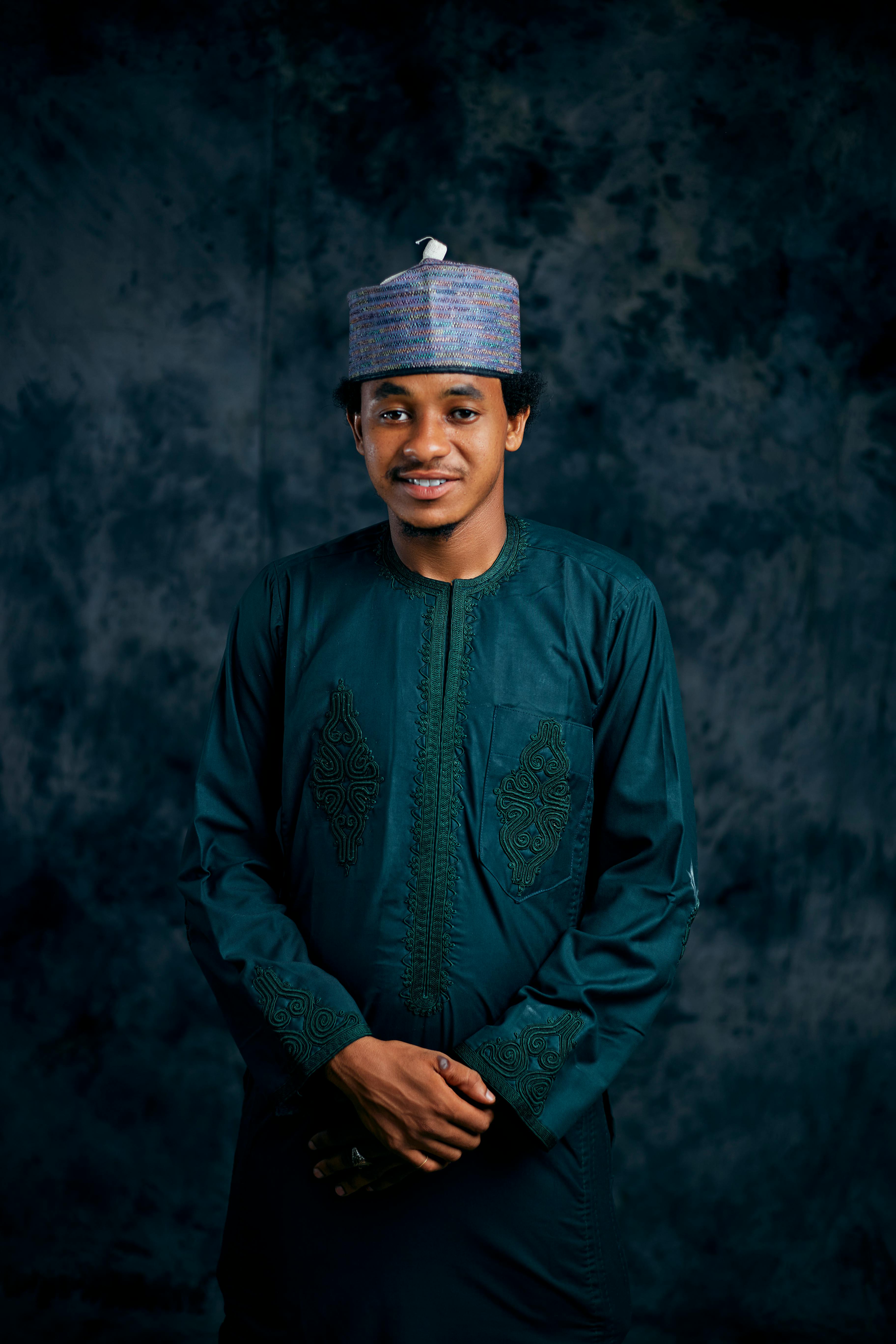 A confident man wearing traditional attire stands against a dark textured background.