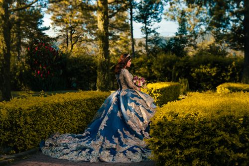 Free A woman in an ornate blue gown stands in a vibrant garden holding a bouquet. Stock Photo