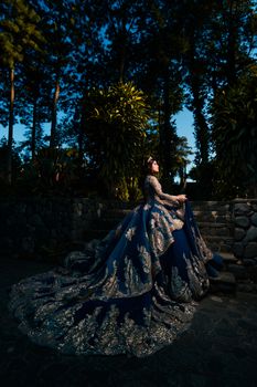 A woman in an elegant gown poses outdoors among trees and stone structures illuminated at twilight.