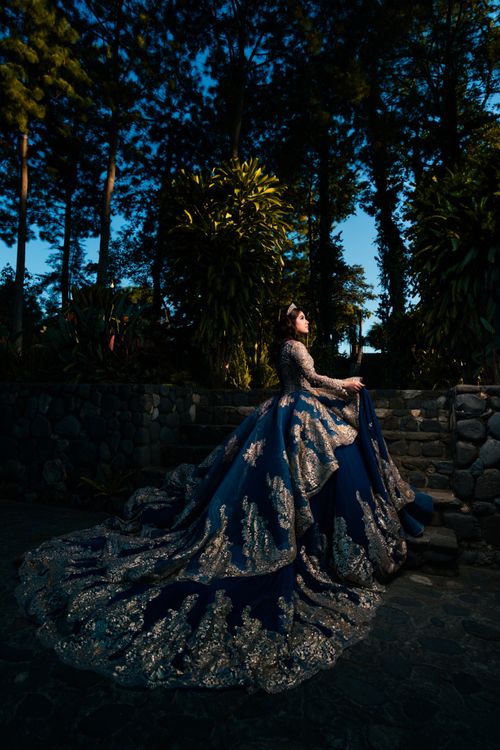 Free A woman in an elegant gown poses outdoors among trees and stone structures illuminated at twilight. Stock Photo