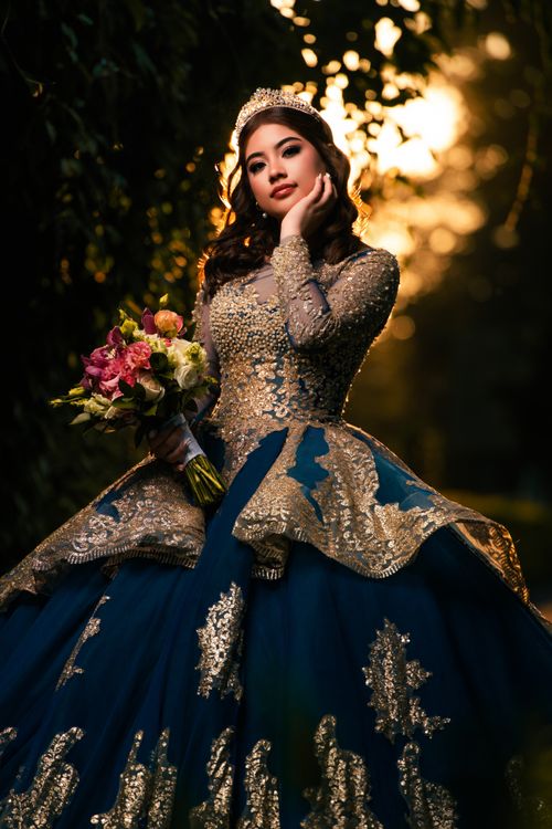 Free Stunning portrait of a woman in a glamorous ballgown, holding a bouquet, against a golden sunset backdrop. Stock Photo