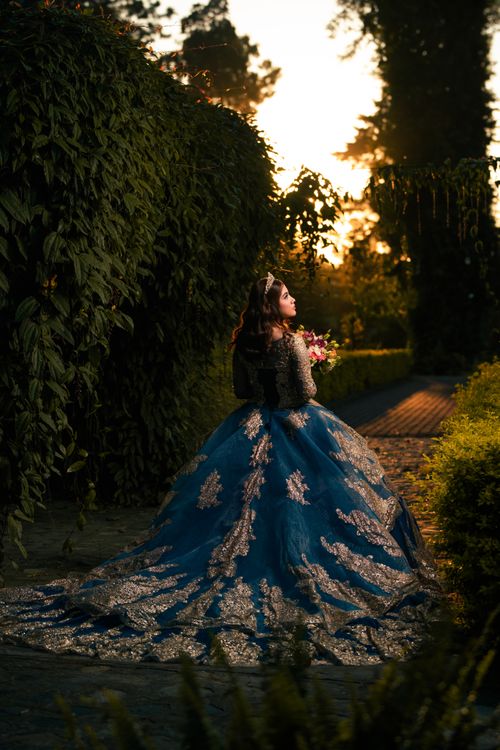 Free A woman in a blue ball gown stands gracefully outdoors, holding flowers at sunset. Stock Photo