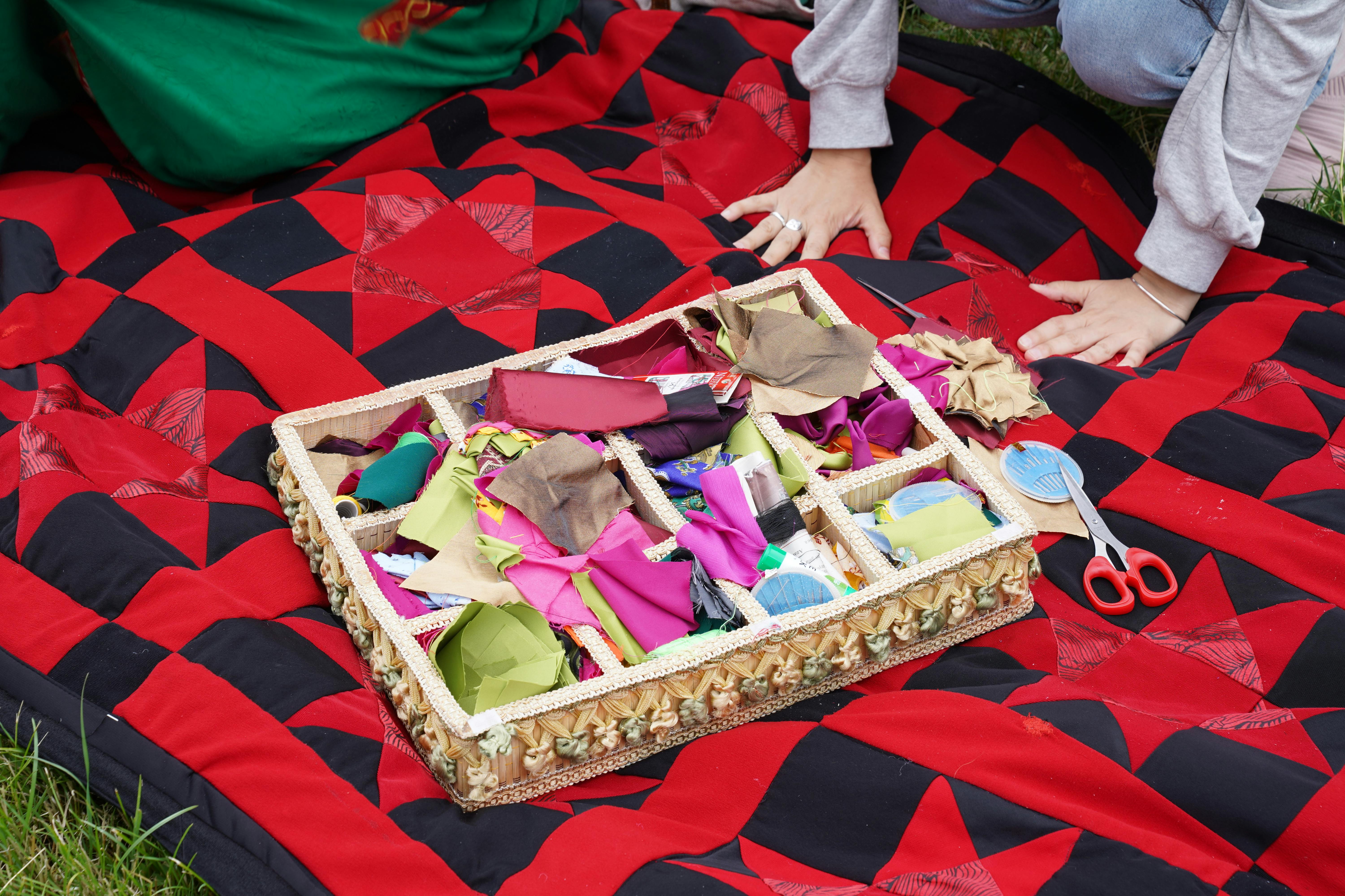 Colorful pieces of fabric and craft supplies arranged outdoors on a red and black picnic blanket.