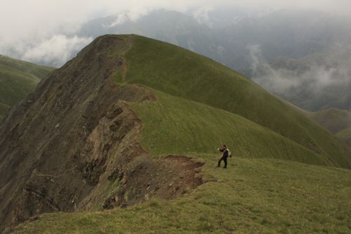 Free Solo hiker navigating steep green mountain ridge under cloudy skies, showcasing adventure. Stock Photo