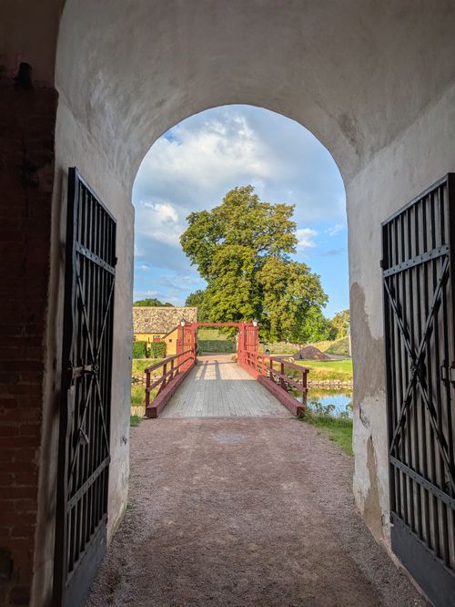 Free A picturesque view of a red wooden bridge captured from an arched stone gate, leading to a lush green park. Stock Photo