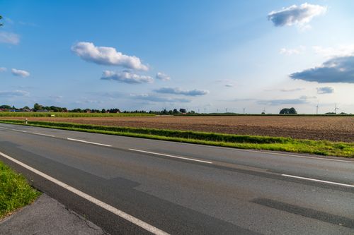 Free A sunny rural scene with an empty road, open fields, and wind turbines in the distance. Stock Photo