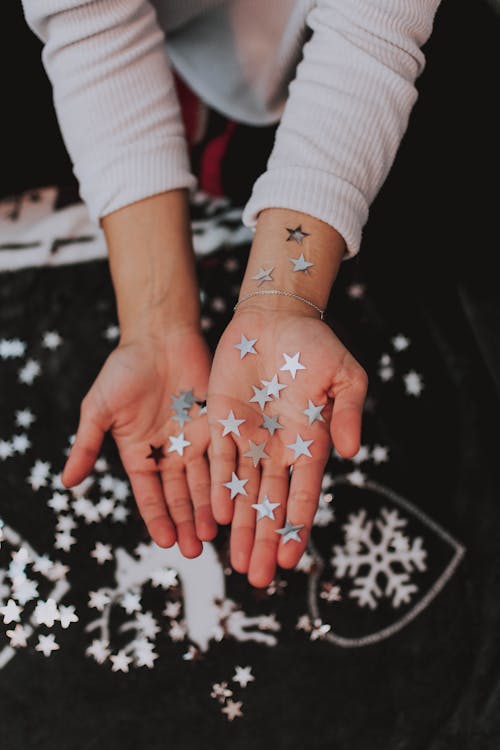 Top view crop faceless female with plenty silver paper stars in hands making cozy Christmas decorations including deer and snowflake placed on black table