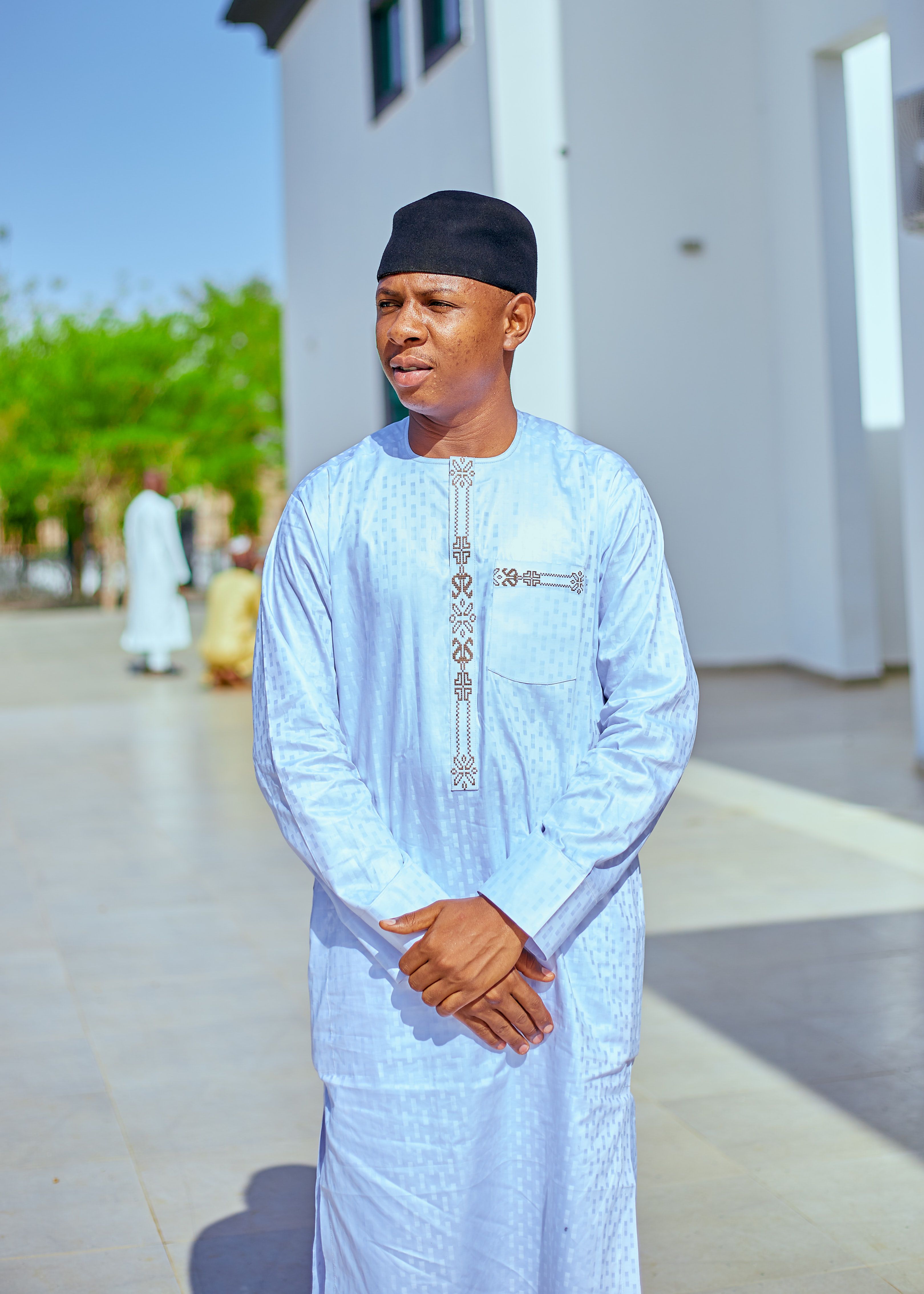 A man in traditional attire standing outdoors, bright daylight, modern white building in background.