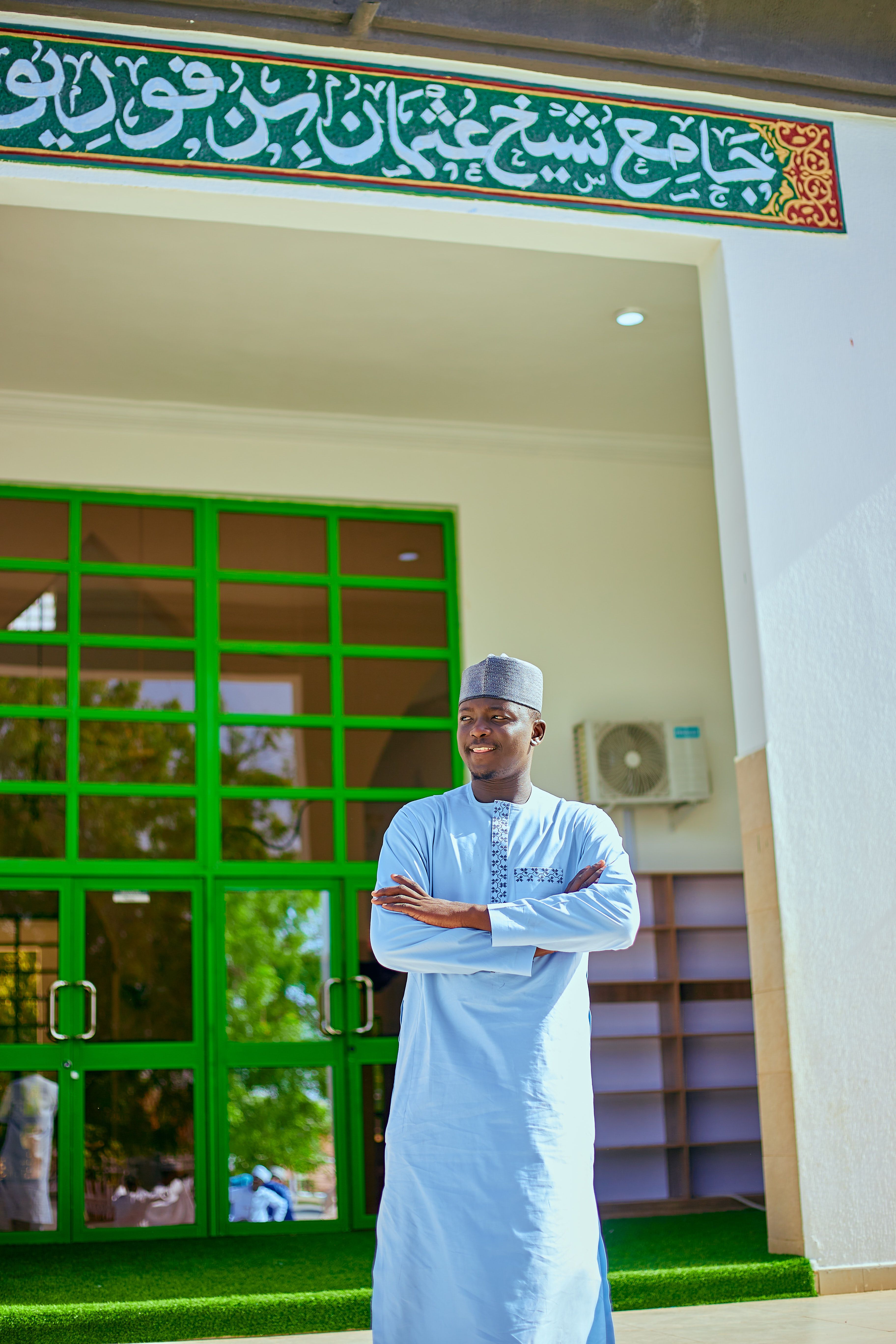 Man poses confidently in front of mosque with green accents and intricate calligraphy.