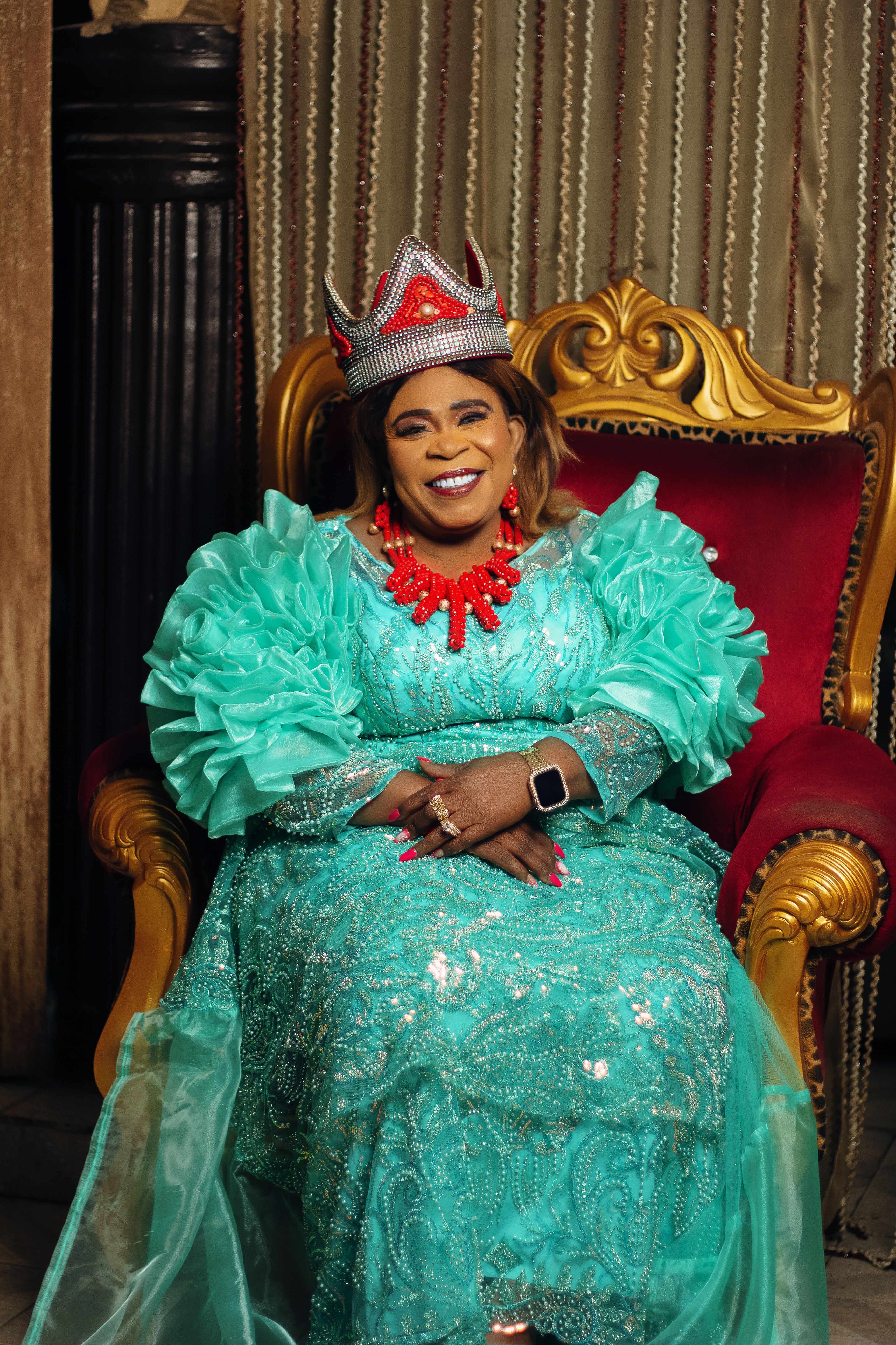 Smiling woman in elaborate turquoise gown and crown, sitting on a regal chair.