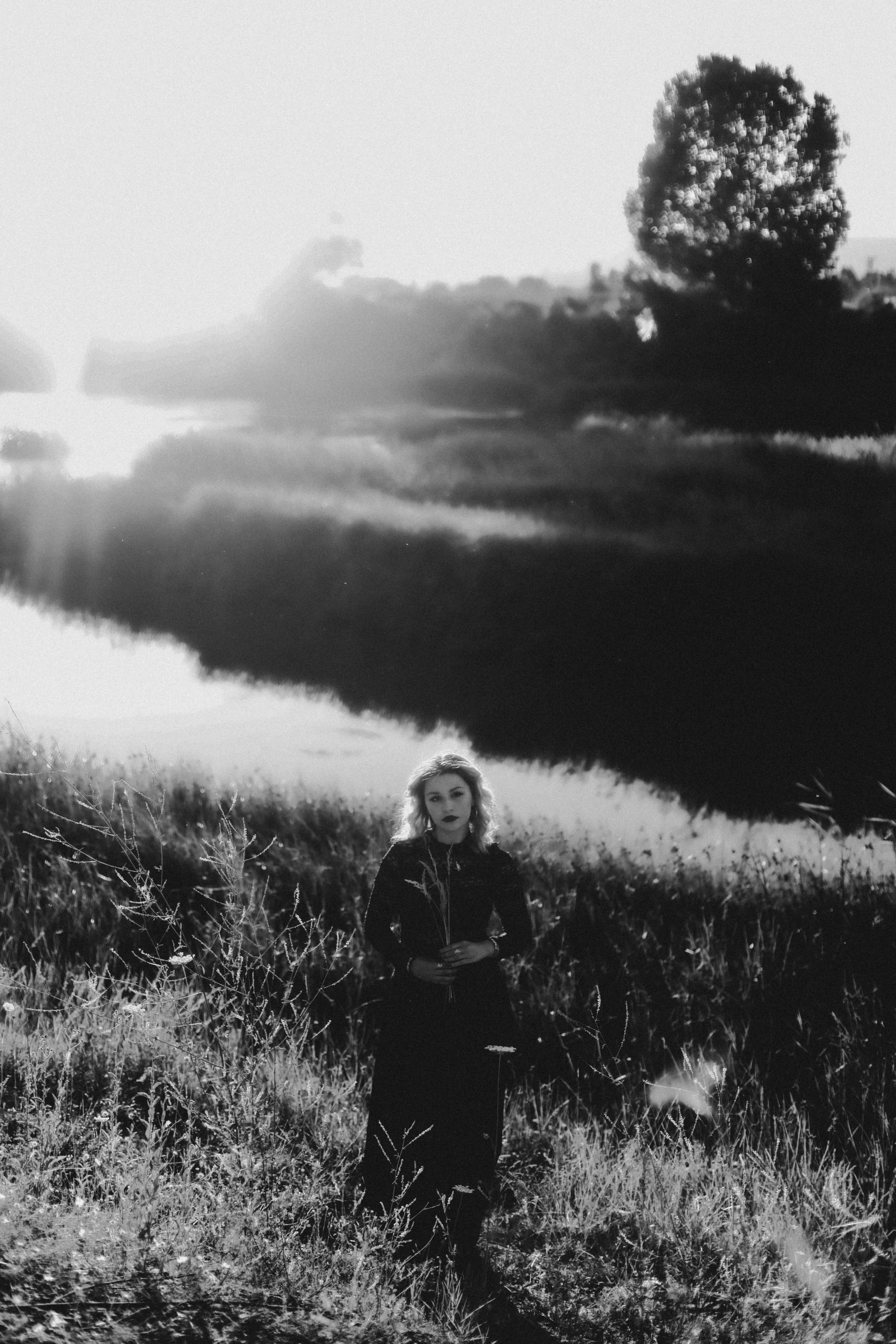 A dramatic black and white portrait of a woman by a serene riverside at sunset.