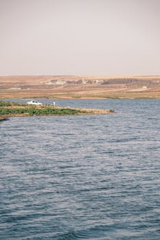 Peaceful riverside scene with a distant vehicle, rolling hills, and serene waters.