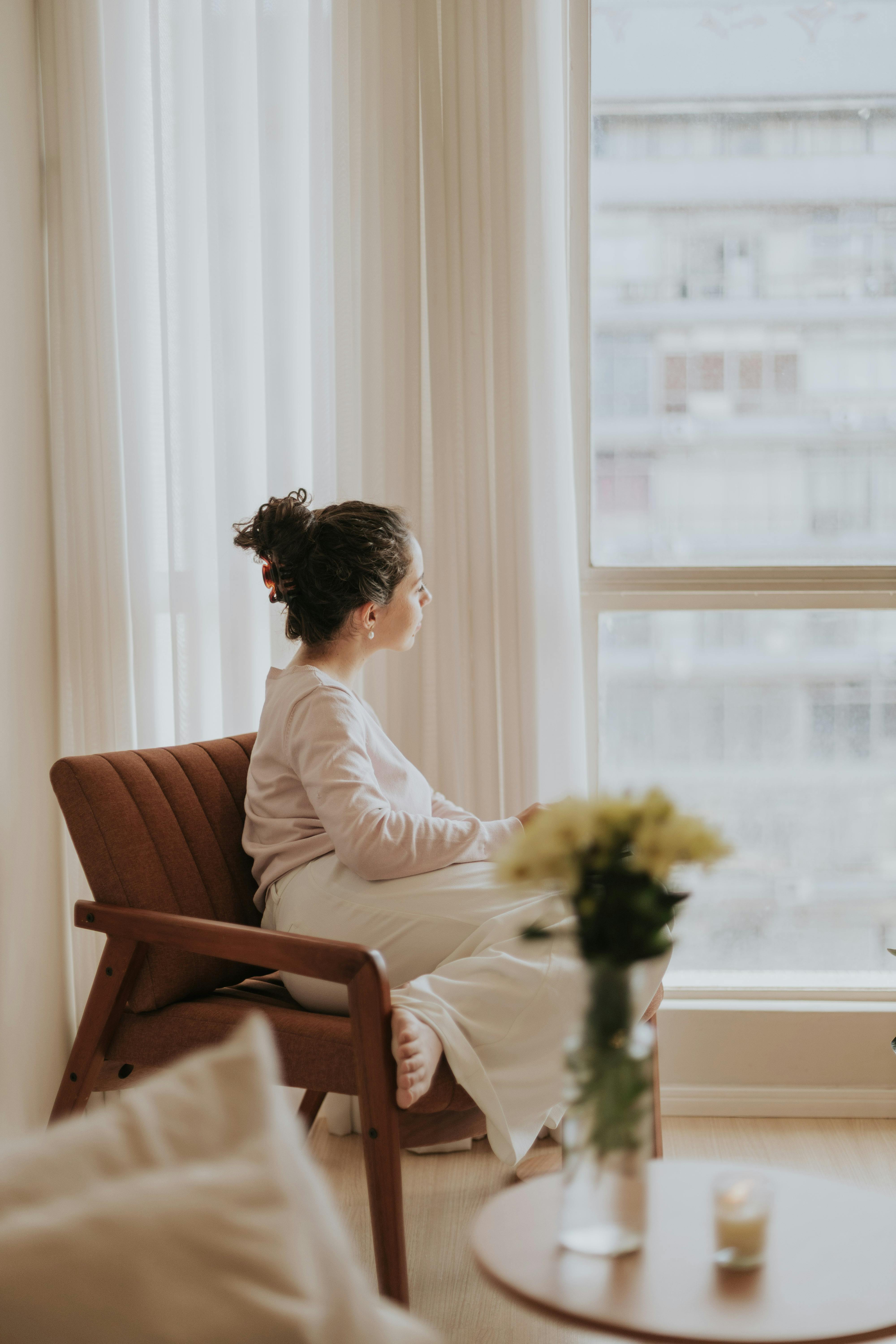 A woman sits in a cozy room gazing thoughtfully out of a large window, surrounded by soft light and warm interiors.