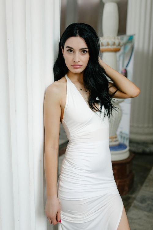 Free Chic portrait of a woman in a white dress leaning against a column indoors. Stock Photo