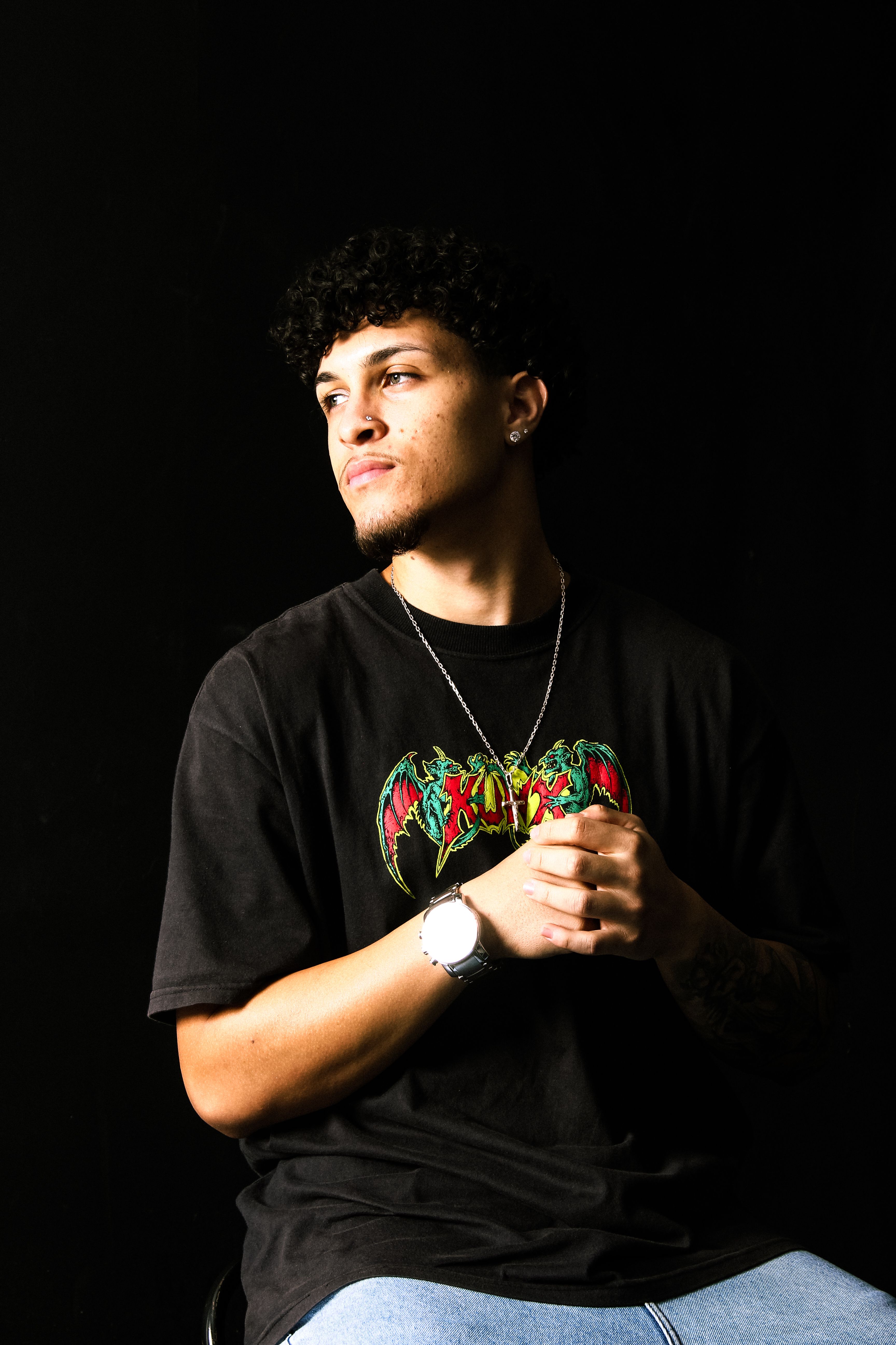 A man in a graphic t-shirt and jewelry poses against a dark background.