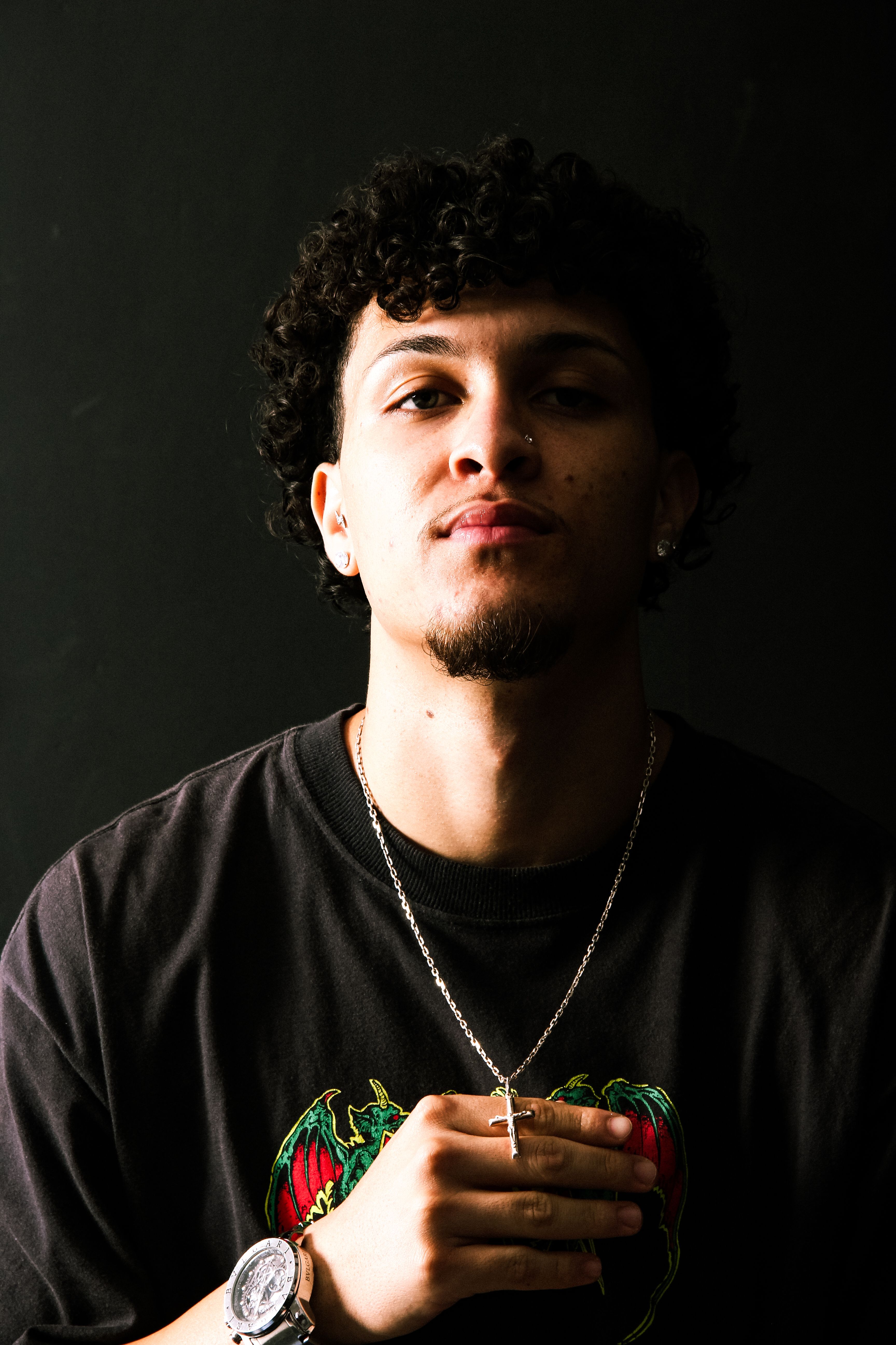 Portrait of a young man with curly hair, wearing a necklace and watch, captured in low-key lighting.