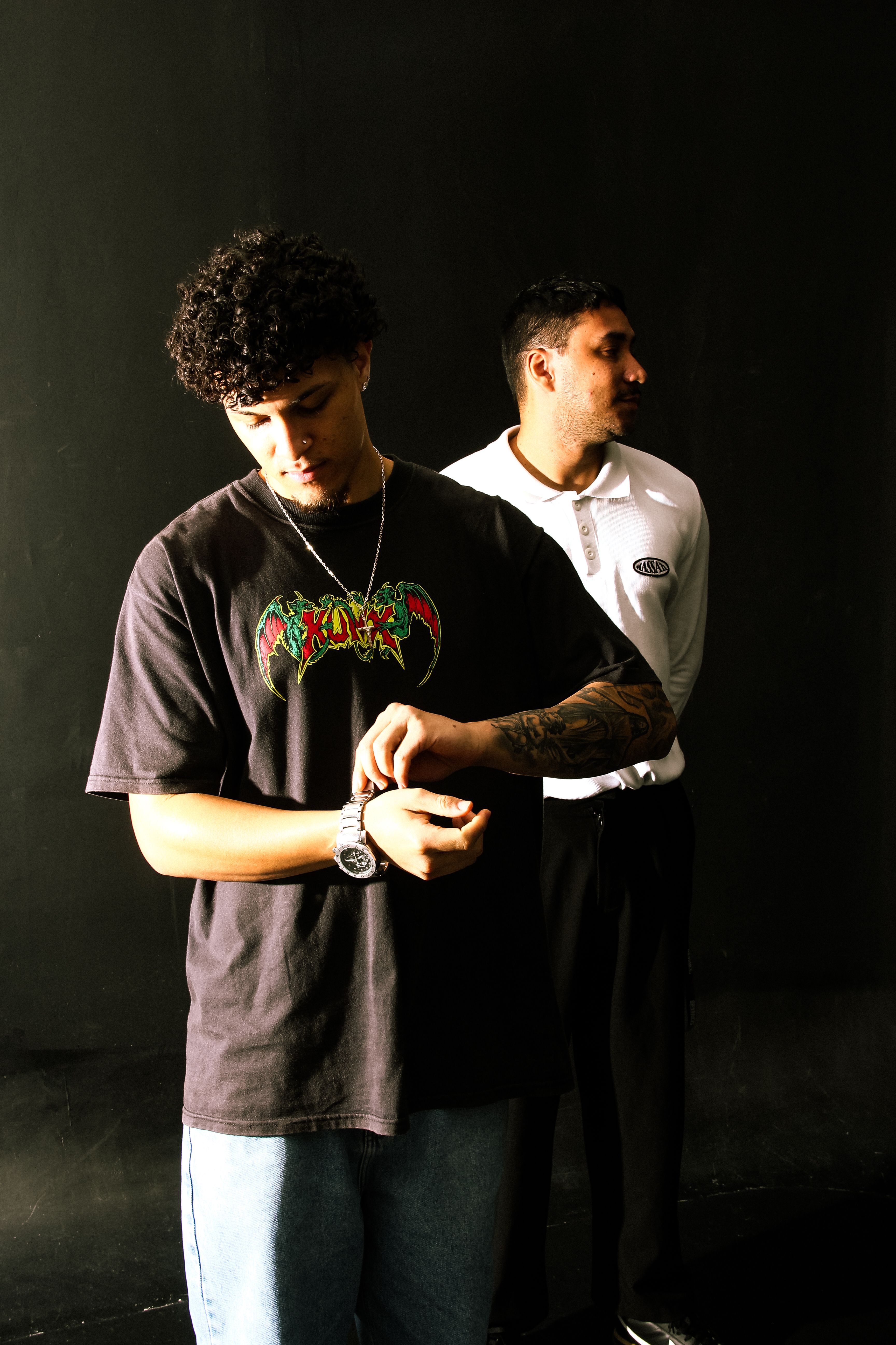 Two young men pose in urban streetwear against a dark backdrop, showcasing modern style.