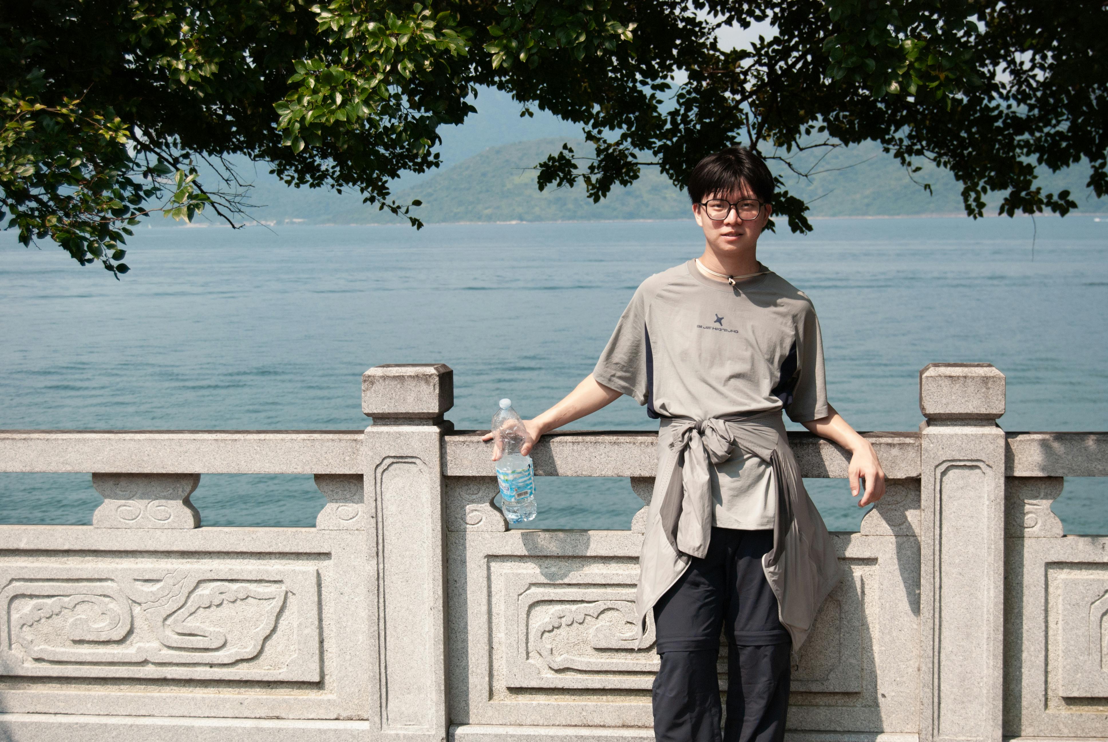 Asian male stands by stone railing holding water bottle with scenic ocean view in Hong Kong.
