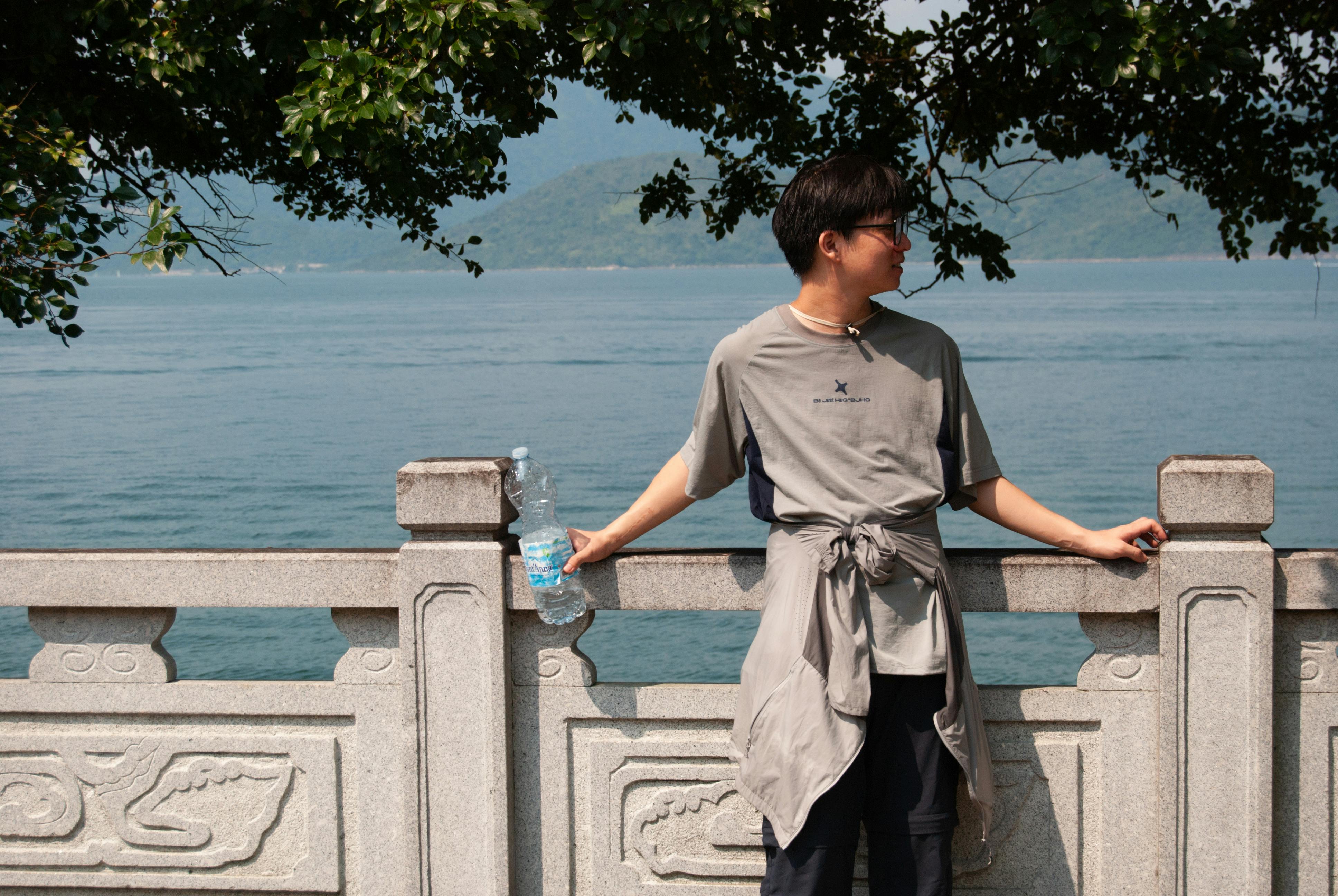 A young man relaxes by the water on a sunny day in Hong Kong, framed by lush trees.