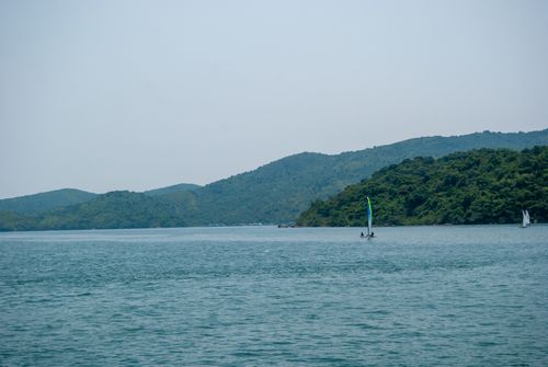 Free A serene view of sailboats on blue waters against lush green rolling hills near Hong Kong. Stock Photo