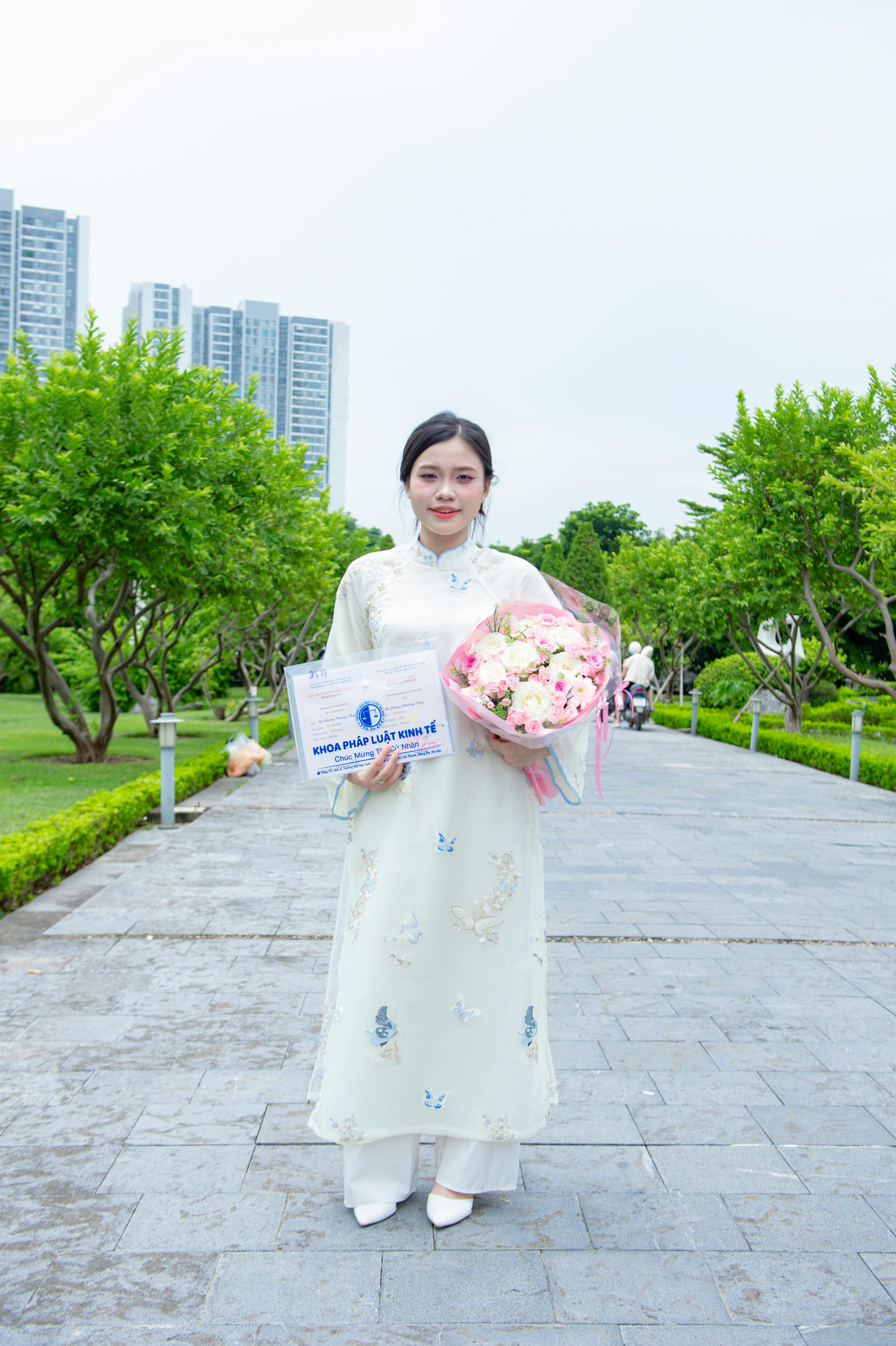 Elegant woman in áo dài standing in Hanoi park with flowers, celebrating an achievement.
