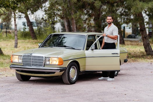 A classic vintage car with its driver standing beside it in an outdoor wooded area.