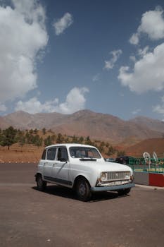 A vintage white car parked in the scenic Moroccan mountains under a partly cloudy sky.