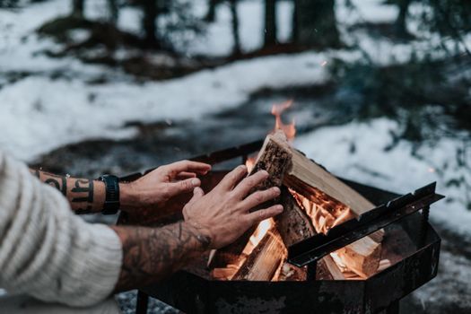 Tattooed hands warming by a campfire in a snowy Lithuanian forest.