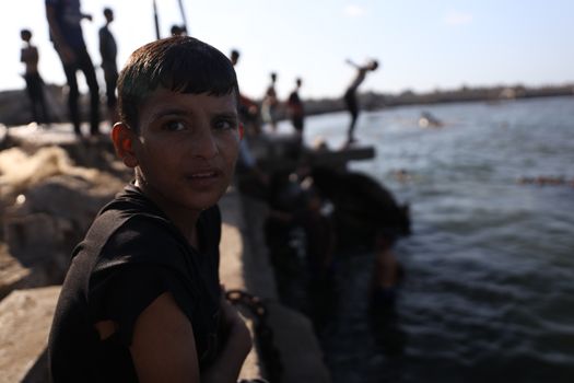 A boy sits by the water as children play, creating a lively scene in Gaza's coastal area.